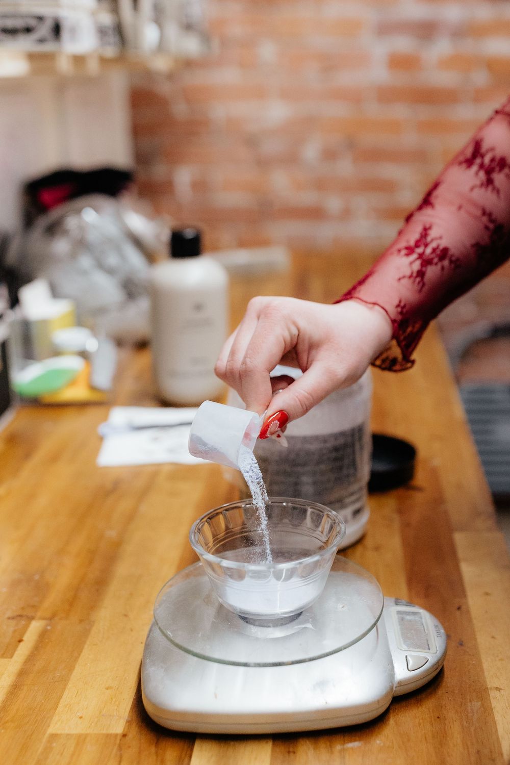 Hand pouring powdered sugar into a measuring scale on wooden butcher block countertop with a brick wall background.