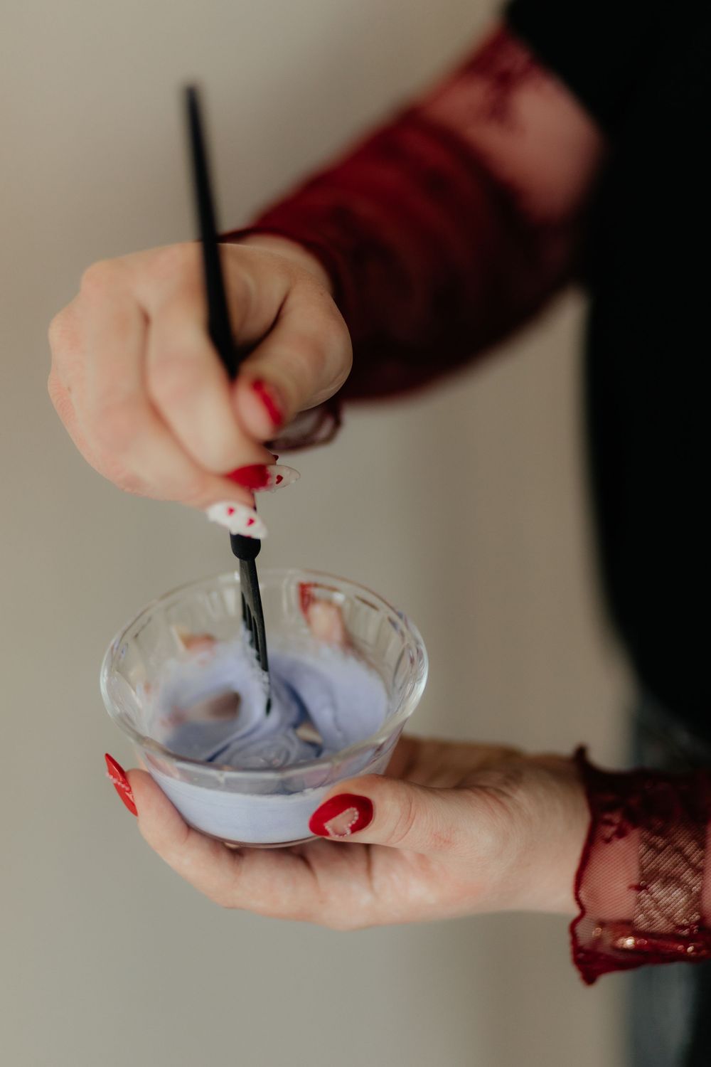 Hands stirring lavender colored mixture in a small glass bowl using a black spoon.