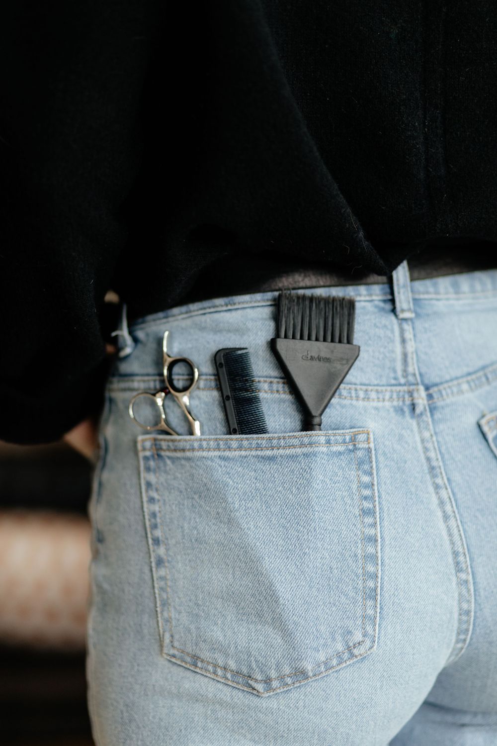 Hair styling tools and clips tucked in the back pocket of light wash denim jeans against a dark background.