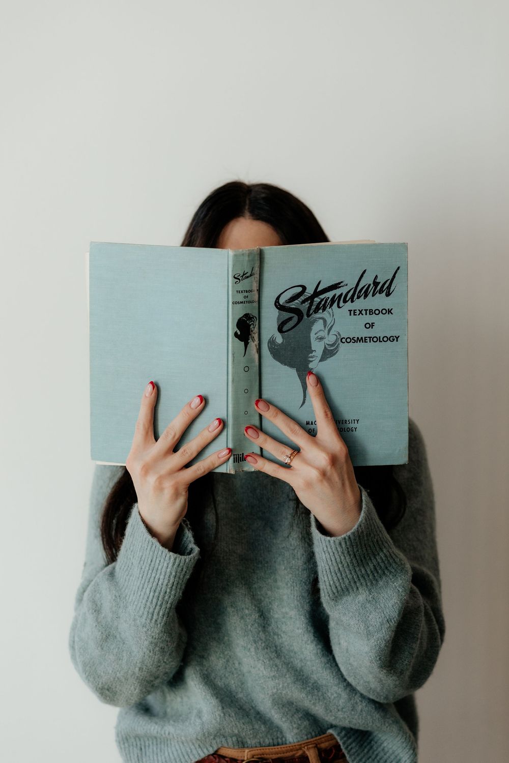 Someone in a gray sweater holds up a light blue book titled 'Standard' covering their face against a white wall.