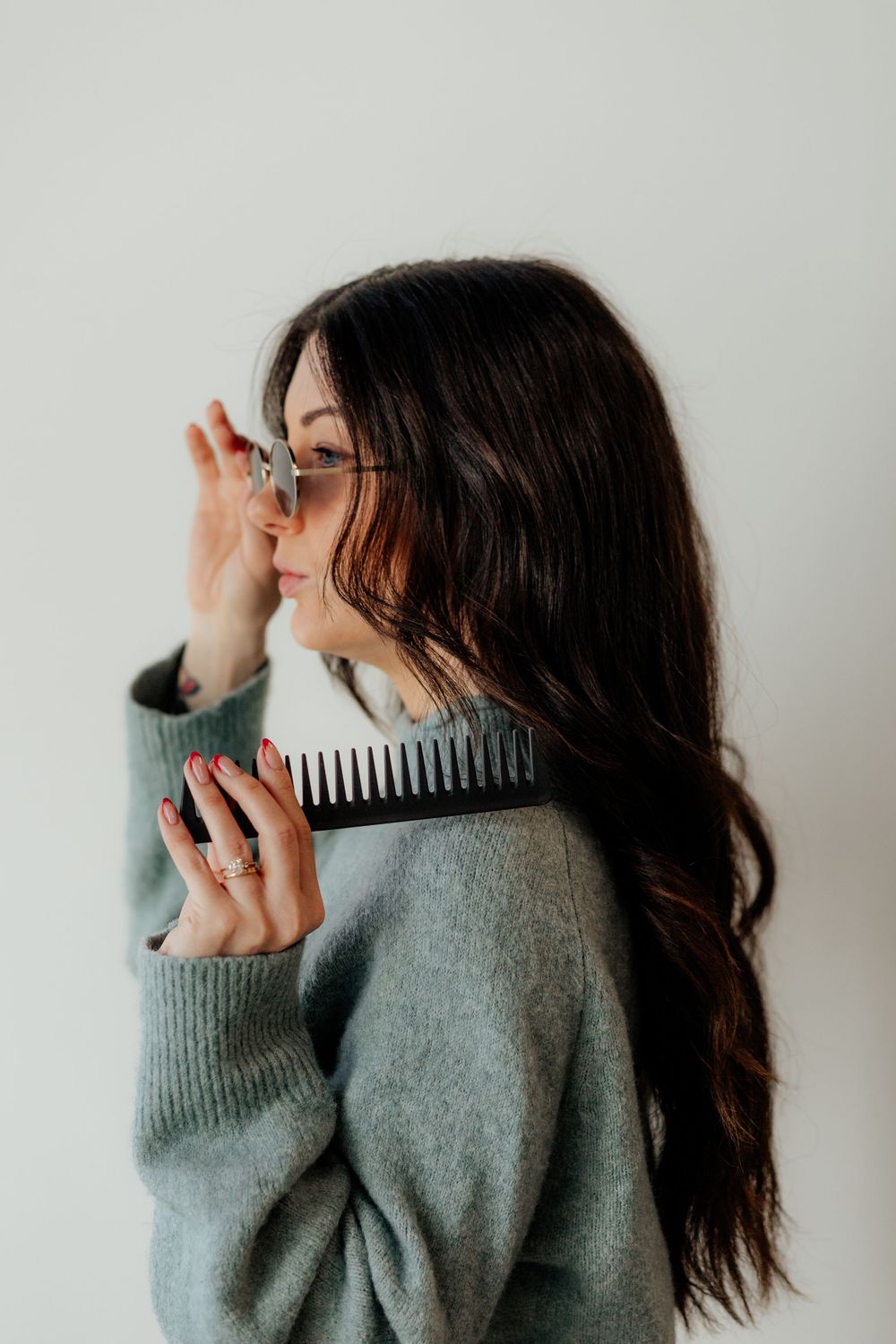 Someone with long dark hair wearing a gray sweater holds a hair comb while wearing glasses against white background.