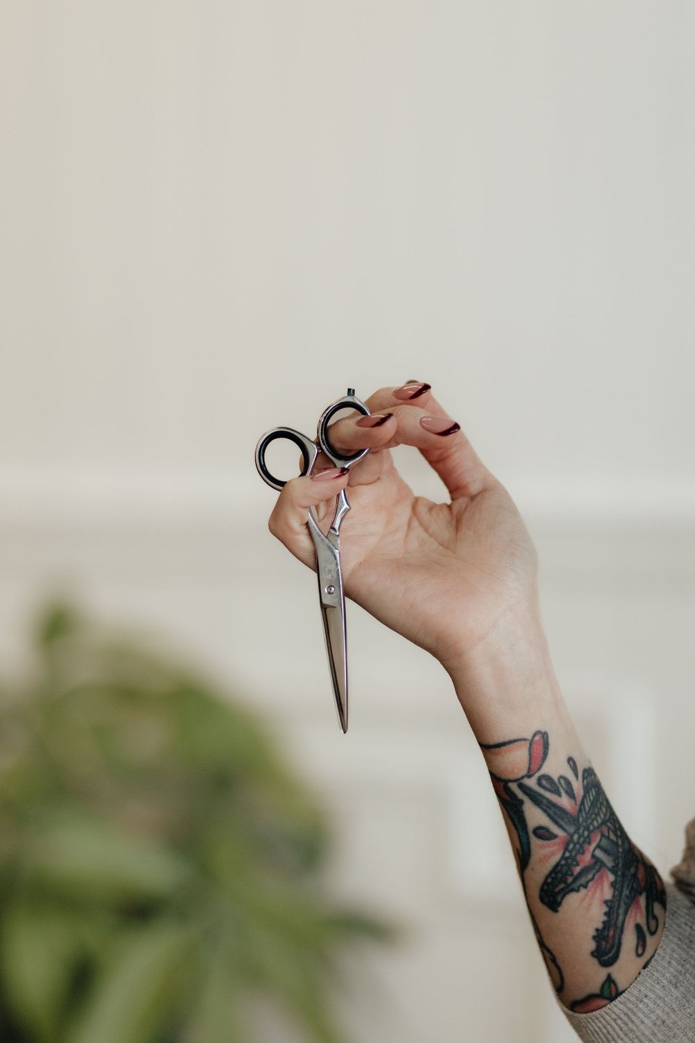 A tattooed hand holds vintage scissors against a light background with green foliage visible in the corner.