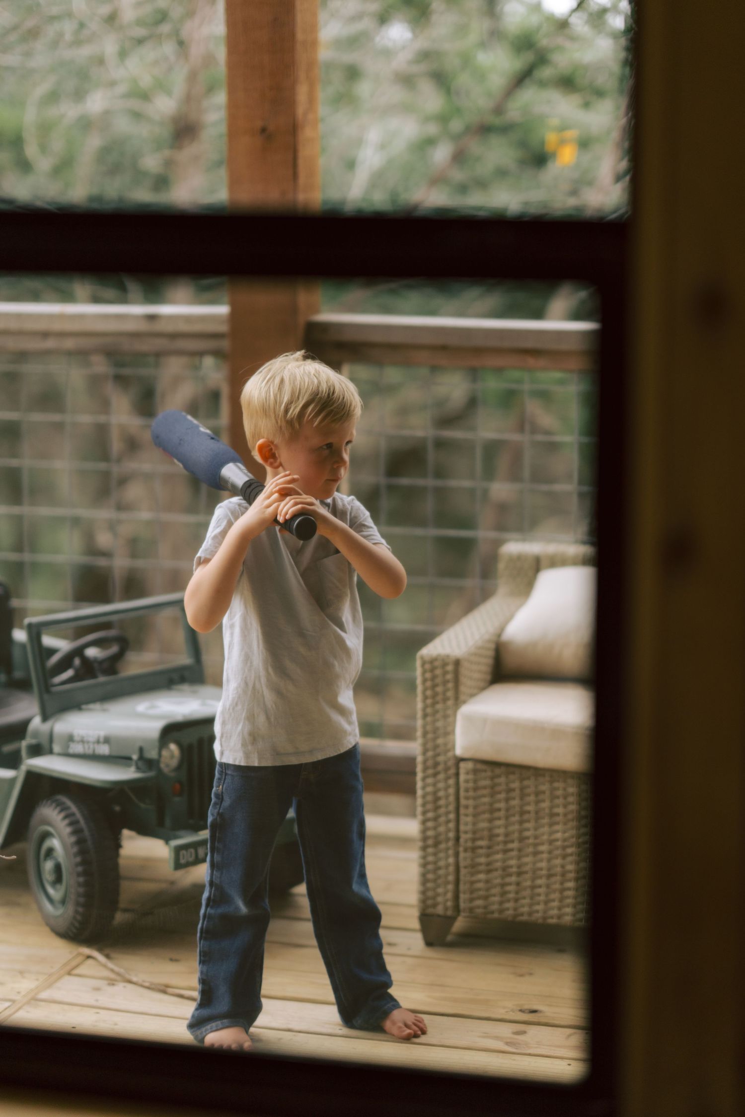 Young child in a white t-shirt plays with a toy on a wooden deck viewed through a window frame.