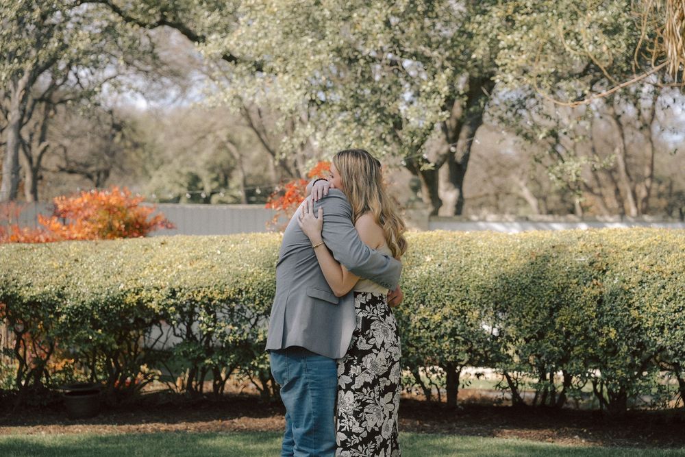 A couple embraces in a serene garden setting with flowering bushes and trees on an autumn afternoon.
