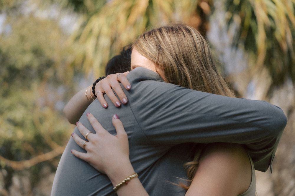 Two people share an intimate embrace outdoors with palm trees visible in the soft-focus background.