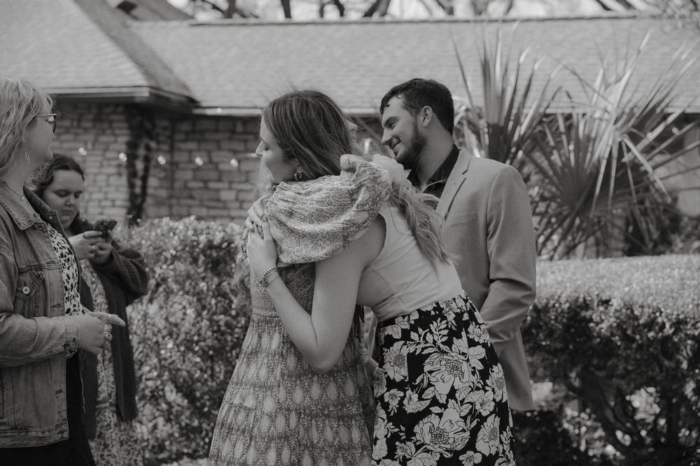 A romantic couple embraces in a garden with palm trees and foliage in the background in black and white.