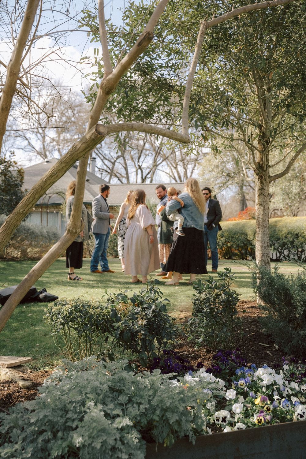 A group gathers in a garden setting with tall trees, blooming flowers, and landscaped grounds on a sunny afternoon.