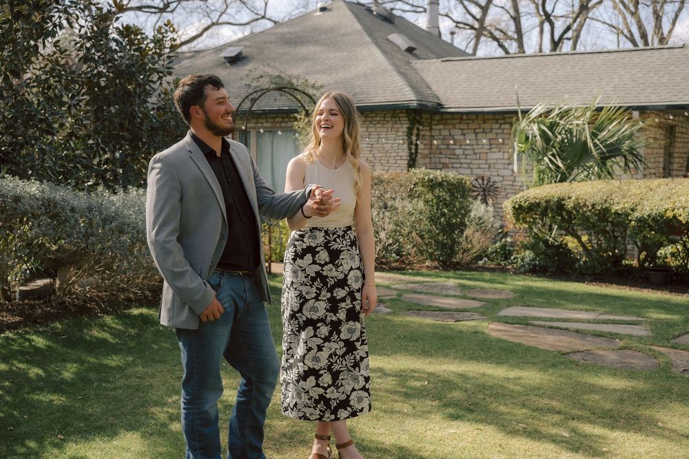 A couple shares a tender moment while standing together on a manicured lawn in front of a stone building.
