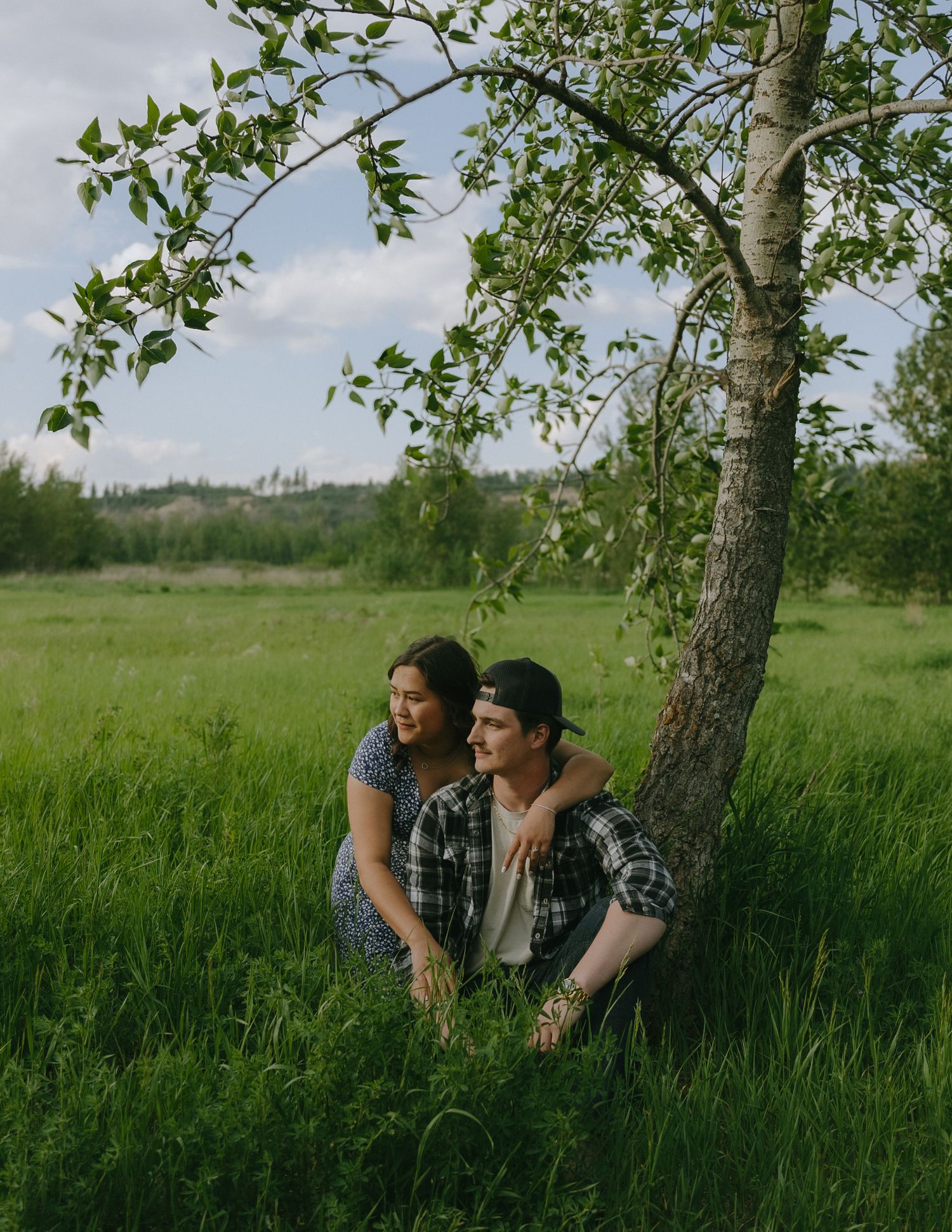 A couple shares a tender moment together under a tree in a lush green meadow on a sunny afternoon.
