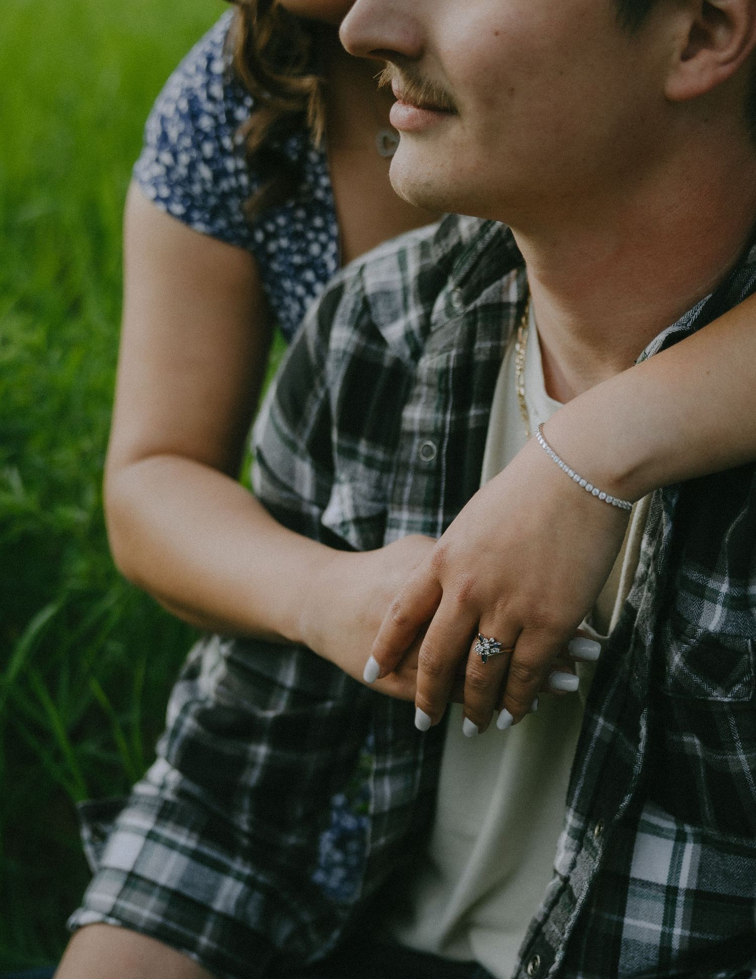 A couple sitting in green grass share an intimate moment as she wraps her arms around him wearing a plaid shirt.