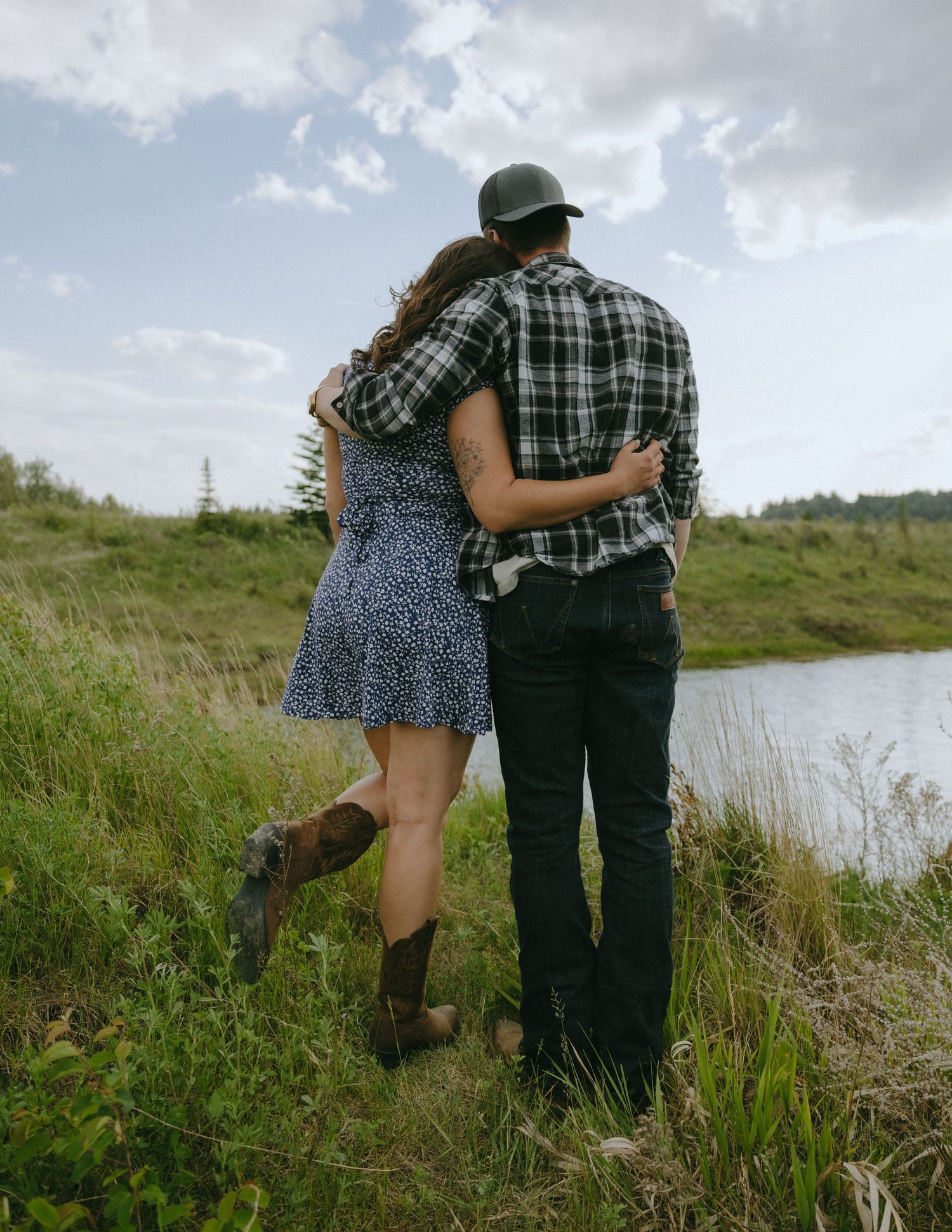 A couple in casual attire embraces while standing near a pond on a cloudy afternoon.