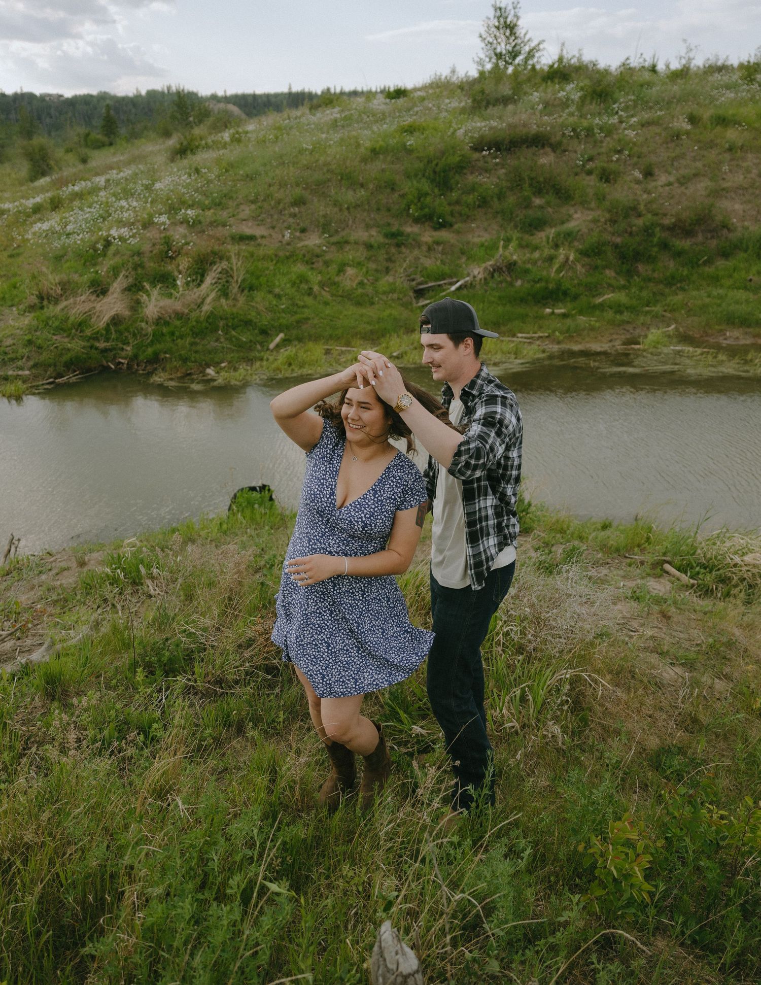 A playful couple dance together on a grassy hillside overlooking a winding river.