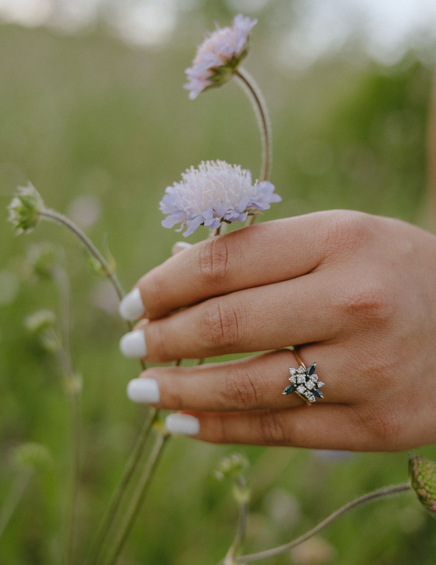 Hand wearing a delicate ring shown against soft purple wildflowers in natural lighting.