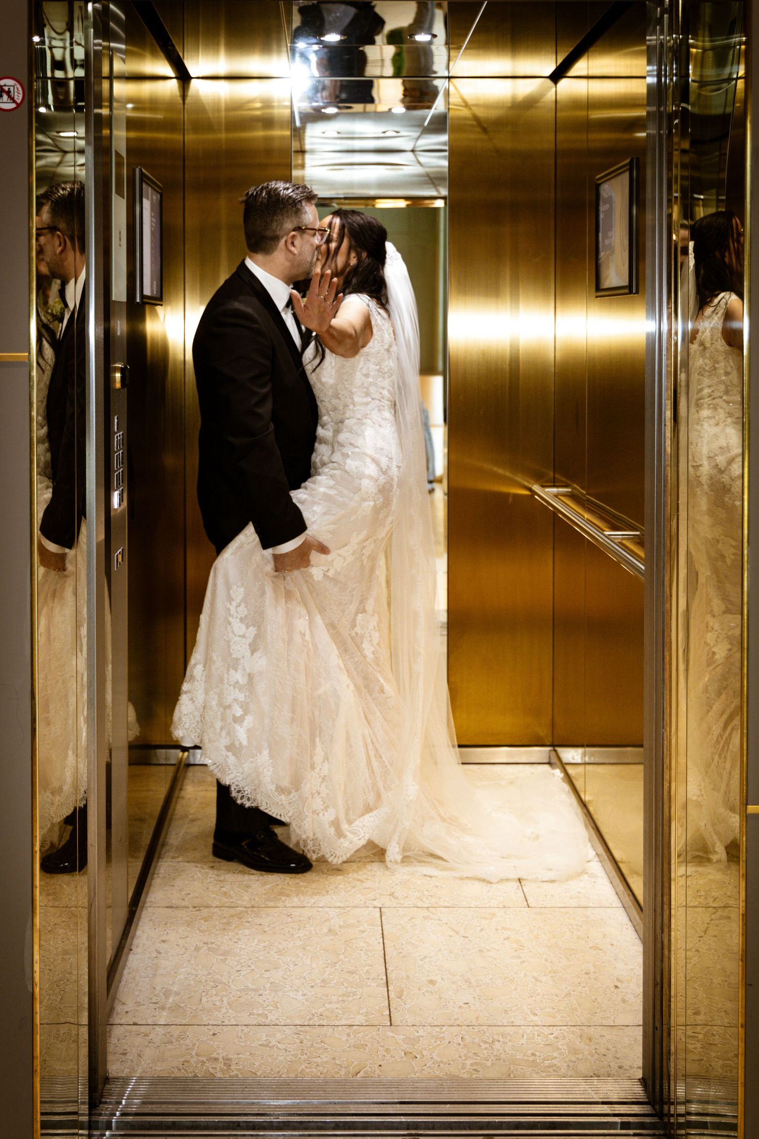 Newlyweds share romantic kiss in gold elevator, bride in lace gown with veil, groom in black tuxedo.