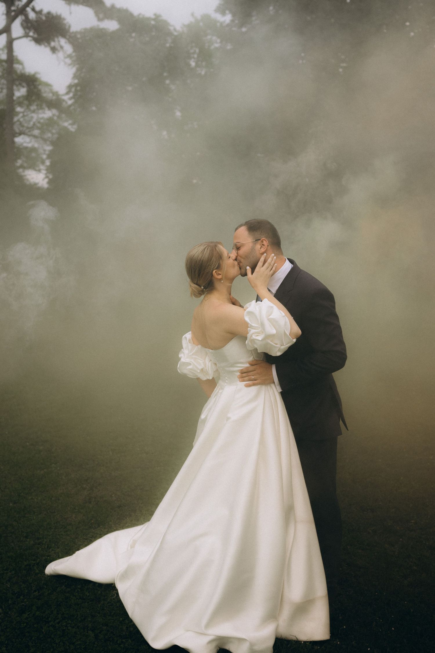 A romantic wedding couple embraces in a dreamy, misty outdoor setting with a white gown and black suit.