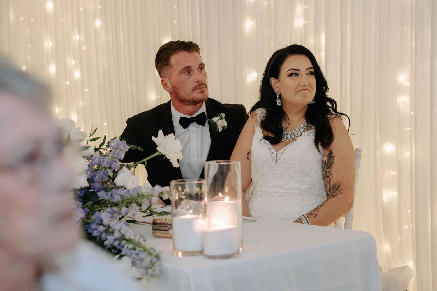 Wedding reception couple shares romantic moment at candlelit table with floral centerpieces and twinkling lights.