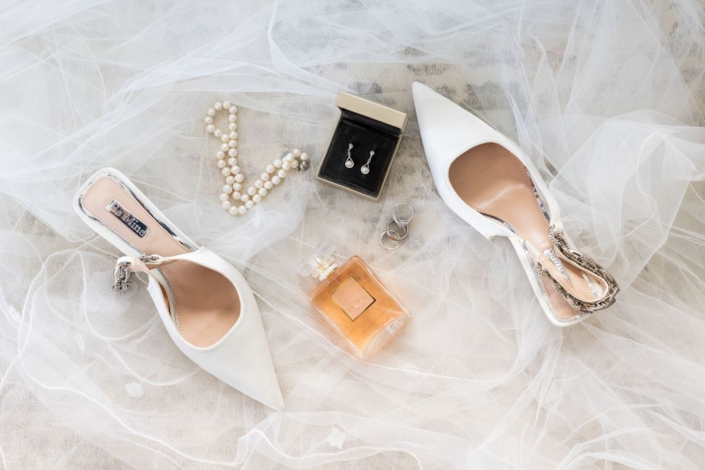 Elegant white bridal shoes arranged with pearl necklace, perfume bottle and ring box on sheer tulle fabric.