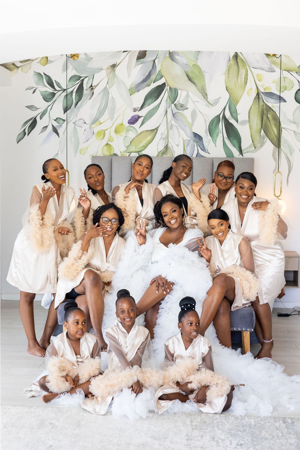 Wedding party group in white robes poses together against floral wall backdrop for pre-ceremony photos.