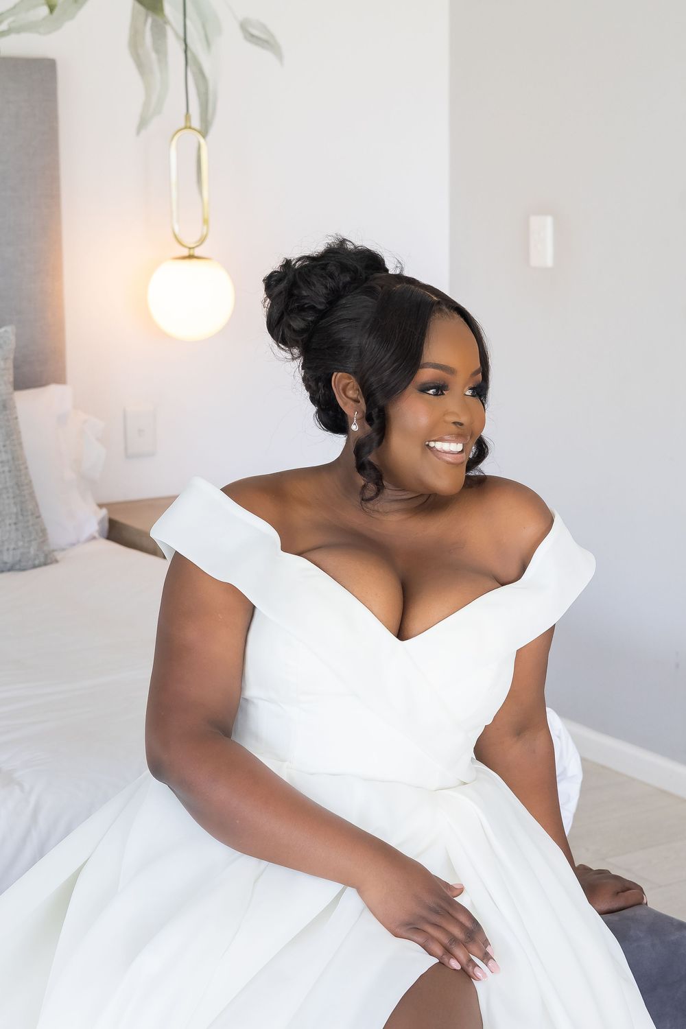 A bride wearing an elegant off-shoulder white wedding gown sits gracefully on a bed under a modern pendant light fixture.