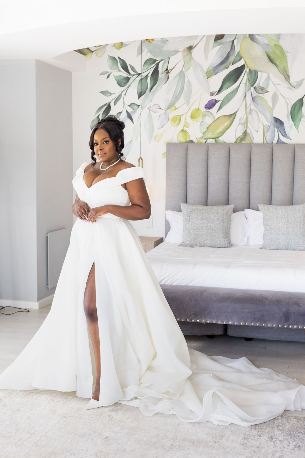 Elegant white wedding dress with off-shoulder design, high slit and long train in modern bedroom with floral wallpaper.
