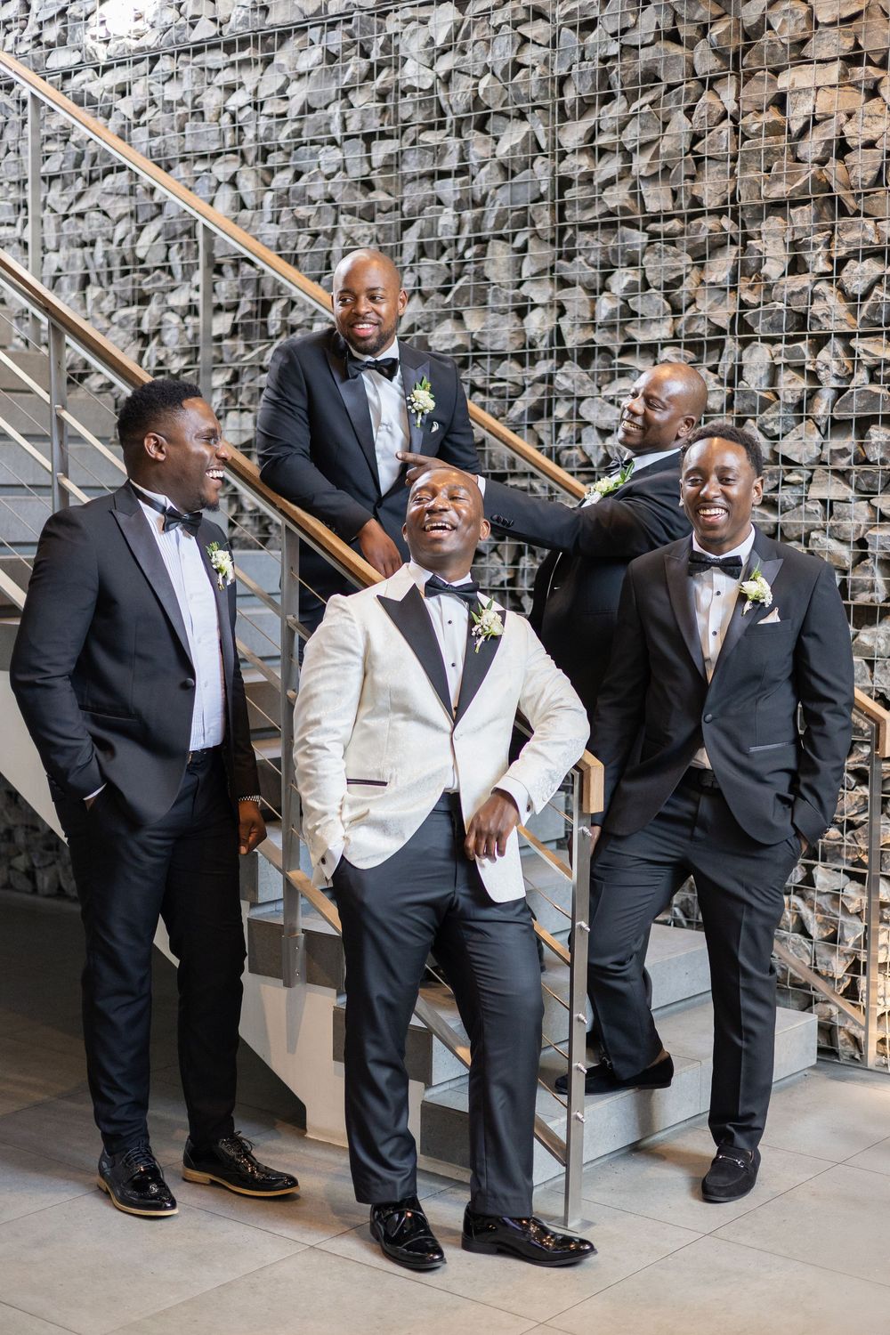 Wedding party in elegant black and white tuxedos pose joyfully on modern concrete staircase with stone wall backdrop.