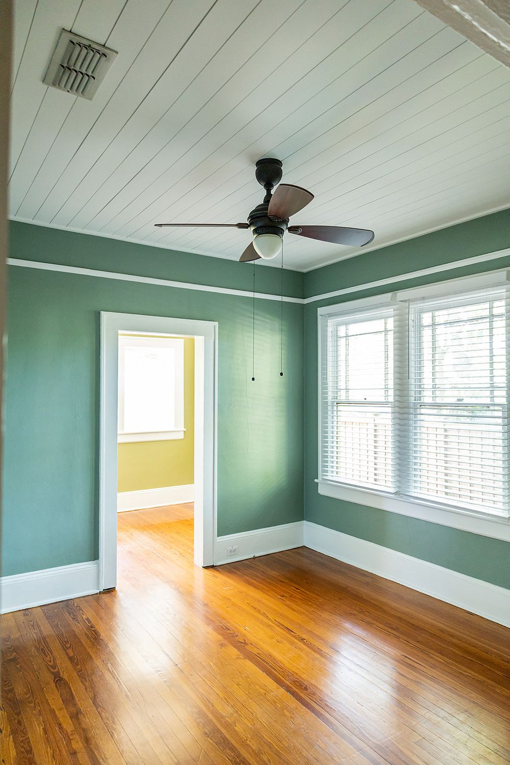 Empty room with mint green walls, hardwood floors, white trim, and a ceiling fan showing doorway to yellow room beyond.