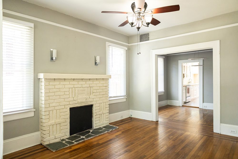 Empty living room features hardwood floors, white brick fireplace, and ceiling fan with natural light from windows.