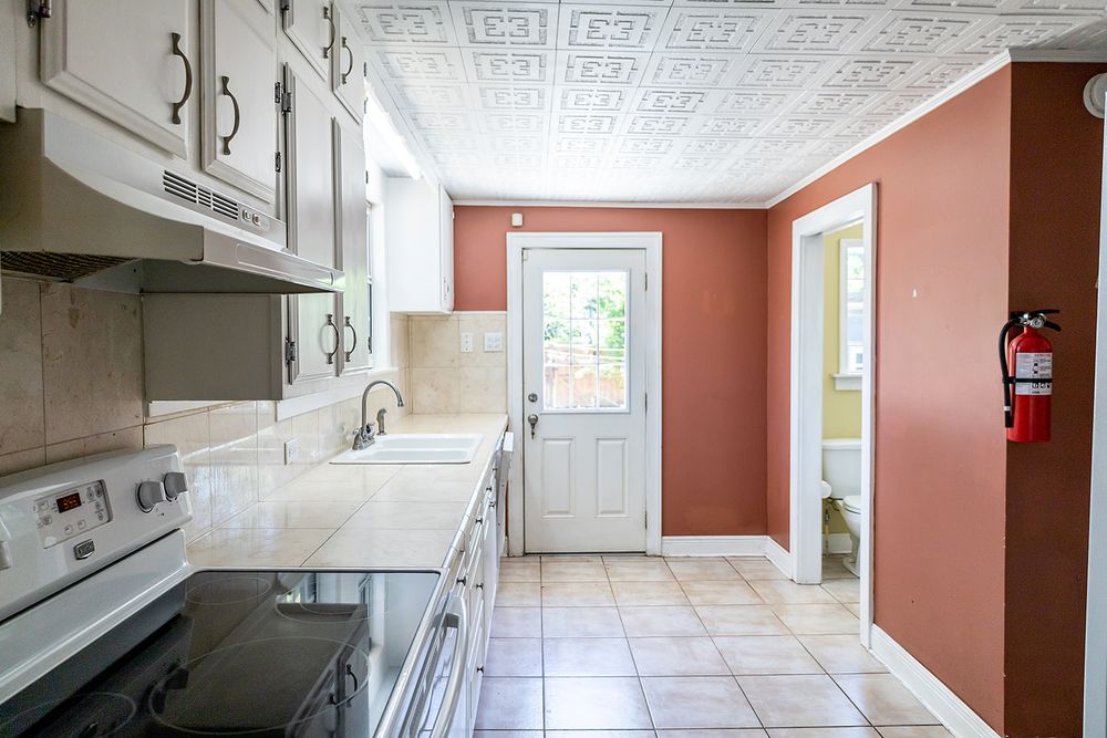 Narrow galley kitchen with white cabinets, terracotta walls, patterned ceiling, and white door leading outside.