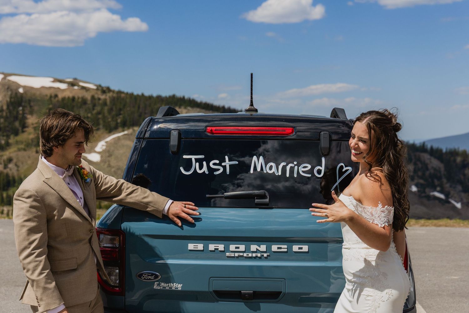 A teal vintage Bronco vehicle parked in front of mountain scenery on what appears to be a wedding day.
