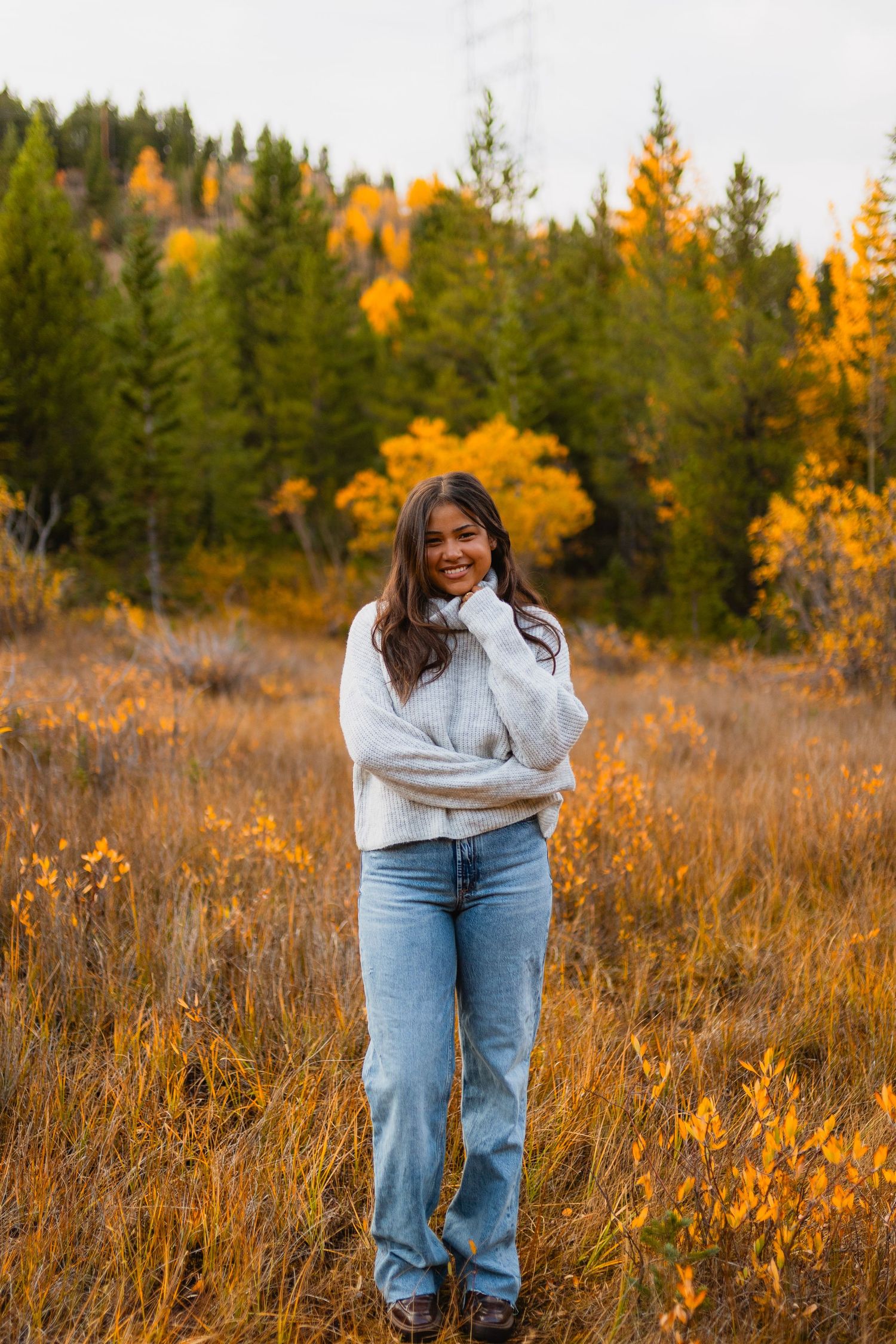 Couple embraces in autumn field surrounded by golden grass and evergreen trees during fall sunset.