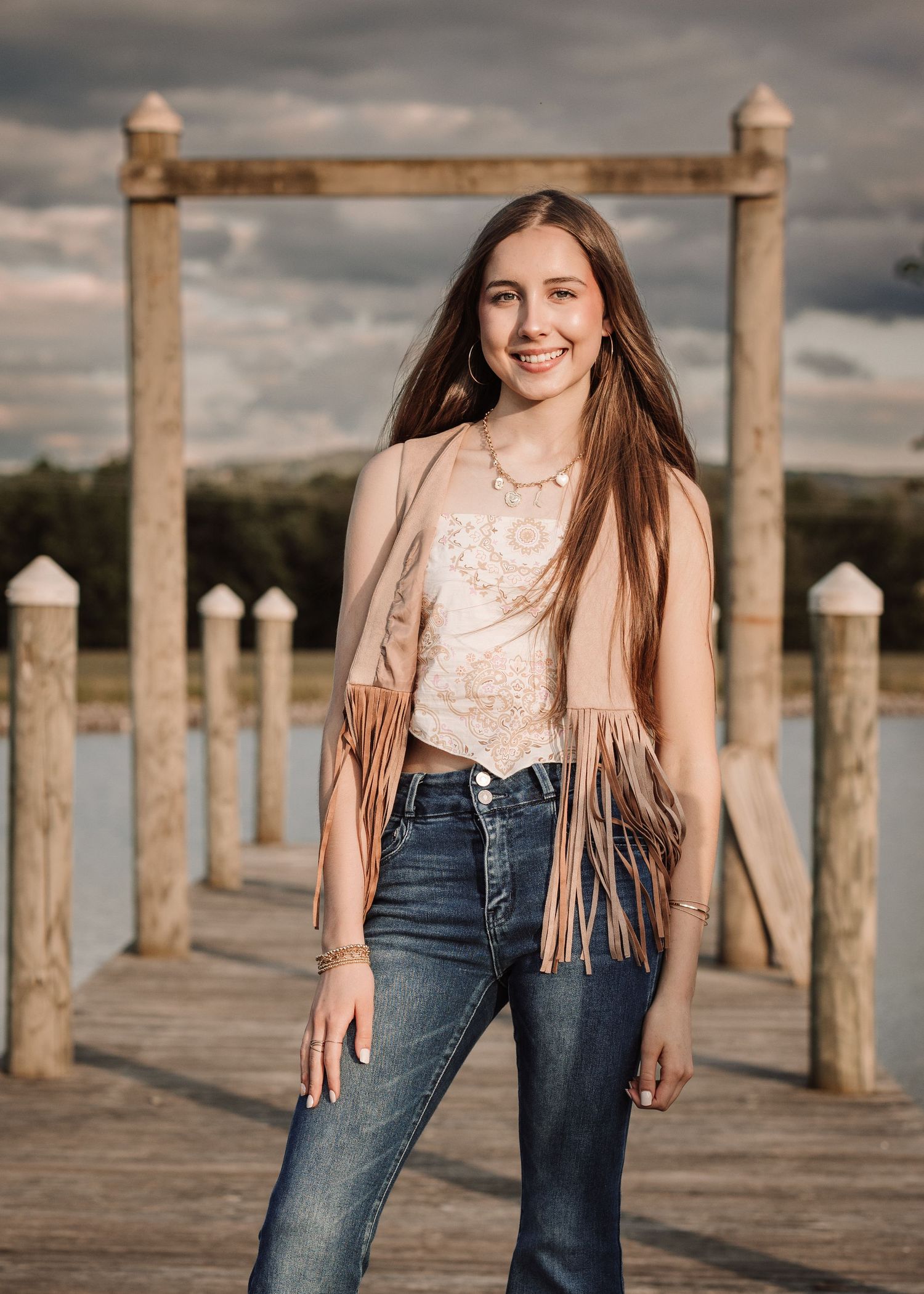 Portrait series on wooden pier with fringe jacket and jeans against dramatic cloudy sky.
