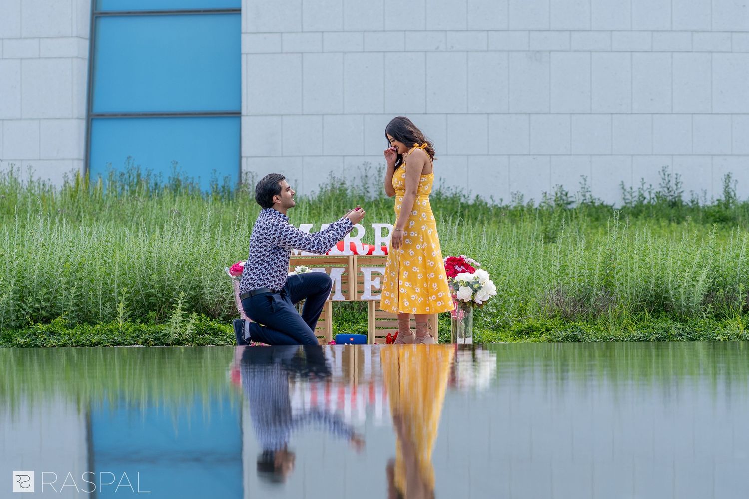 A proposal scene reflected in still water with a blue building and green grass in the background.