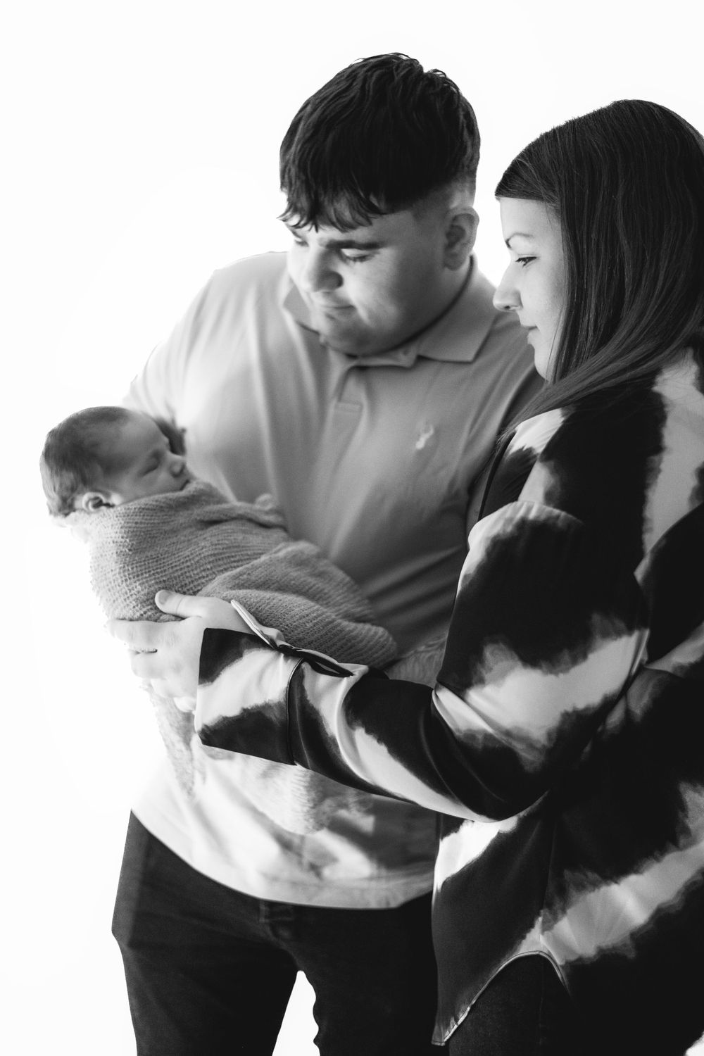 Black and white portrait of parents admiring their wrapped newborn baby against white backdrop.