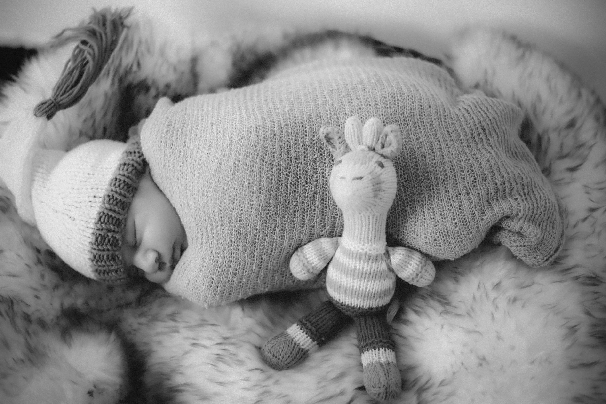 Black and white image of sleeping baby with knit toy on plush surface.
