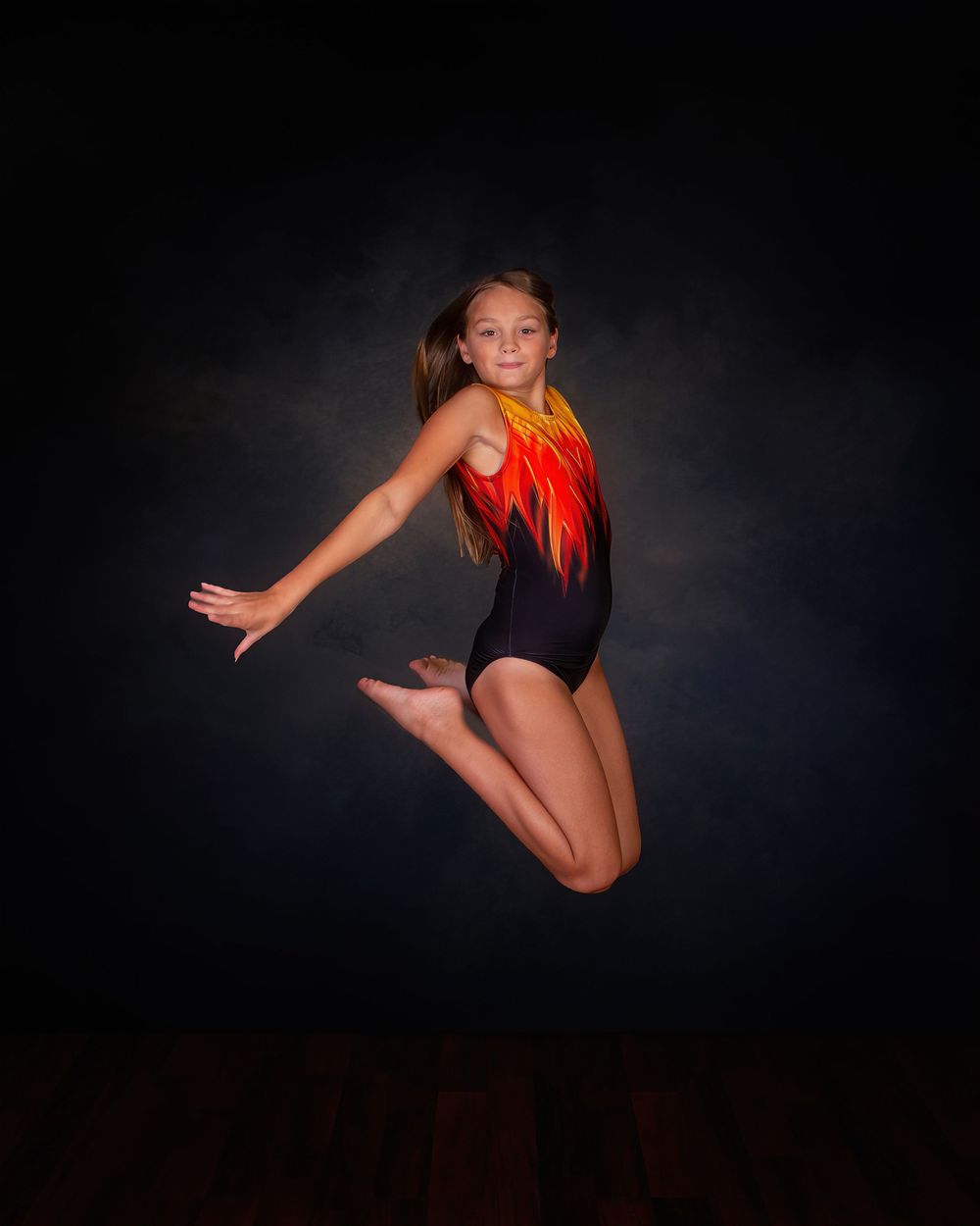 A gymnast in a black leotard with orange accents performs a dynamic jumping pose against a dark dramatic background.