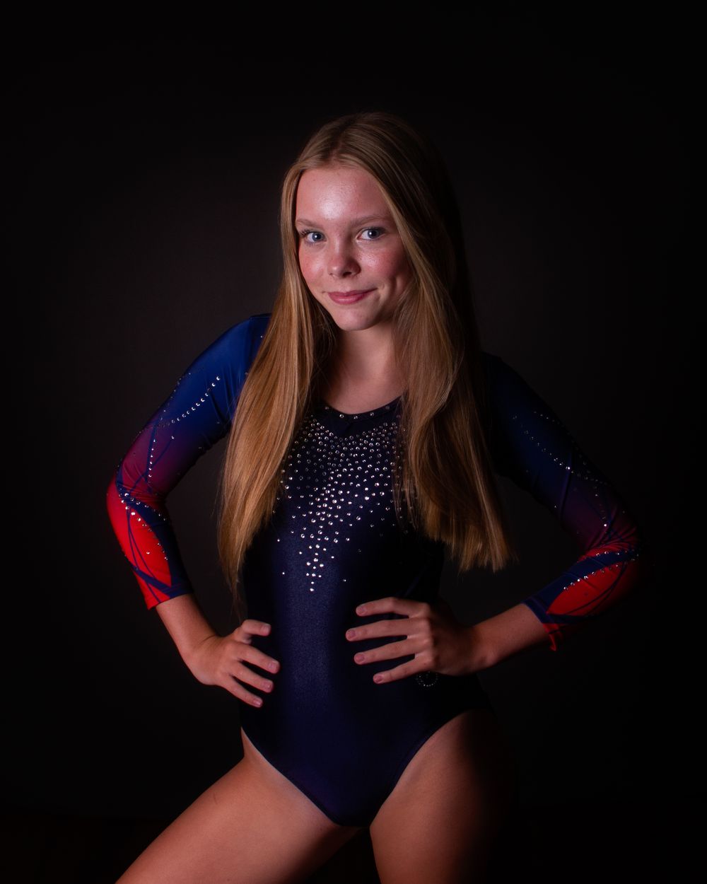 Gymnast in a sparkly navy blue and red leotard poses confidently against dark background during portrait session.