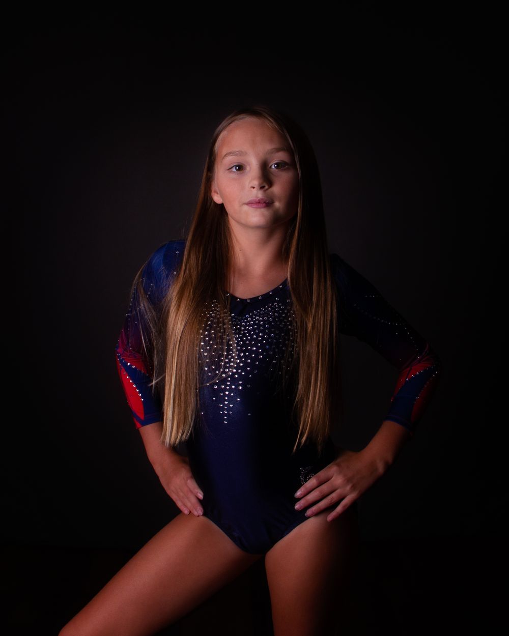 Gymnast strikes a confident pose wearing a navy blue sparkly leotard with red accents against a dark backdrop.