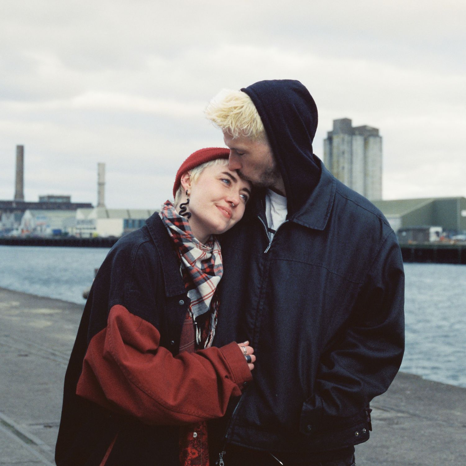 Two people in winter coats embrace tenderly by a waterfront on an overcast day.