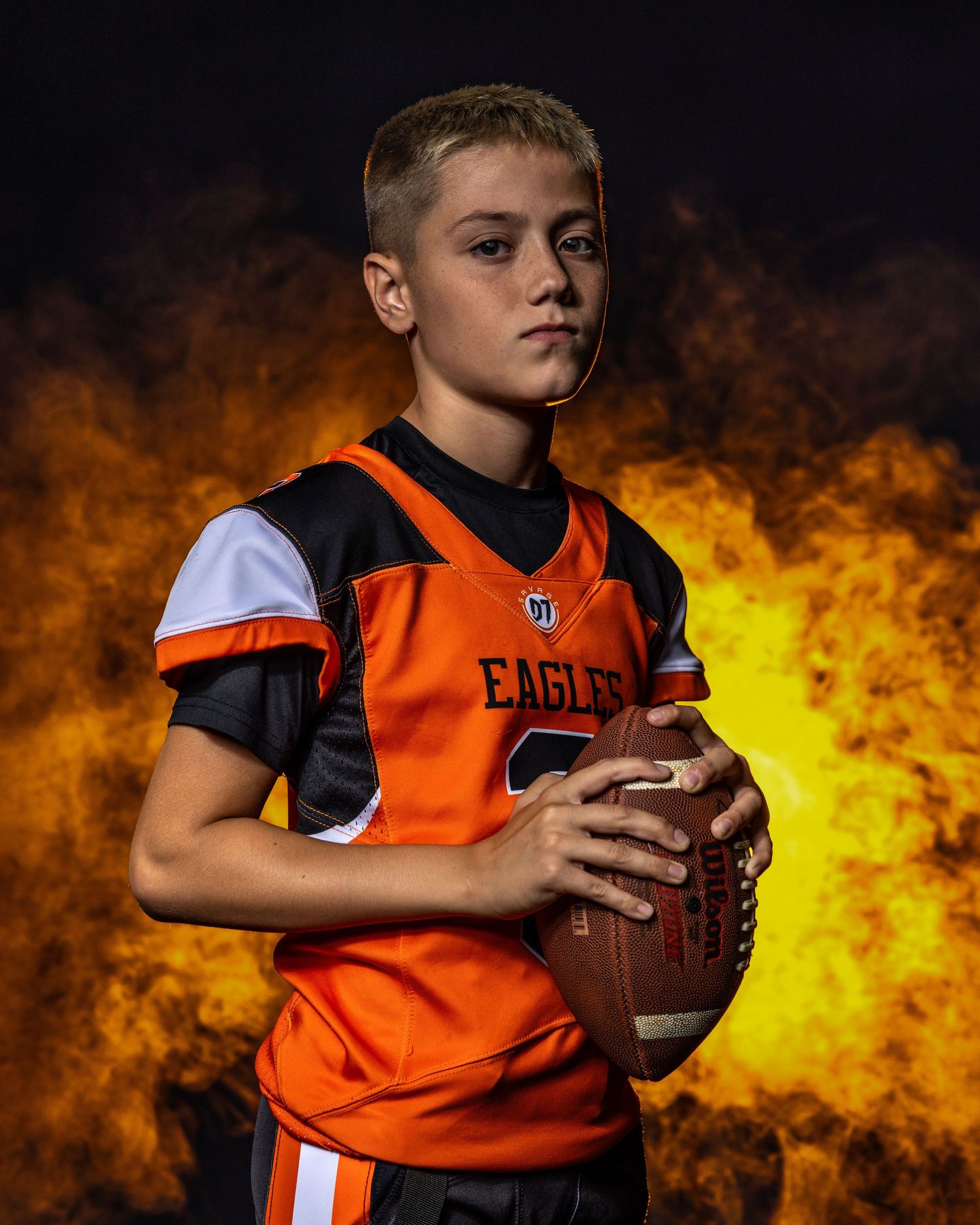 Youth football player poses in orange uniform against fiery background effects.