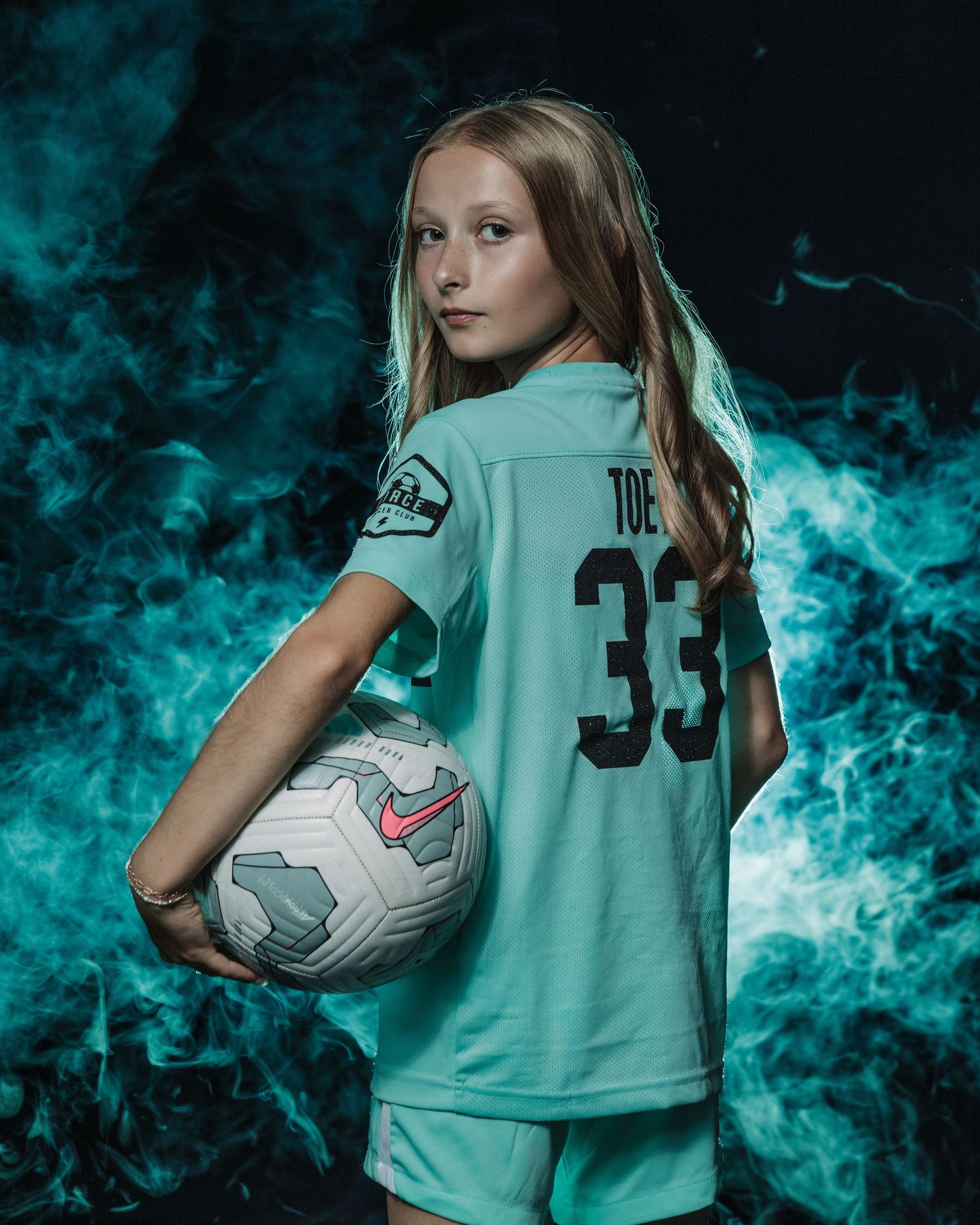 Soccer player poses with ball against teal smoke effect background.