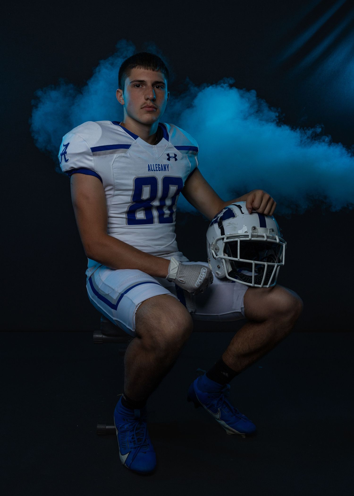 Football player in white and blue uniform poses with helmet against dramatic blue smoke background.