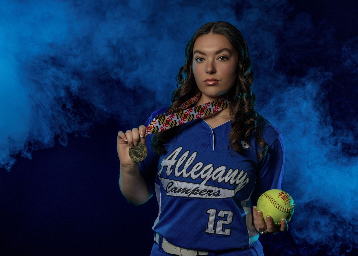 Softball player in blue jersey poses against dramatic dark blue background.