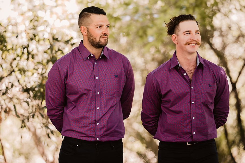Two people wearing matching burgundy dress shirts smile together outdoors in a sunlit natural setting.