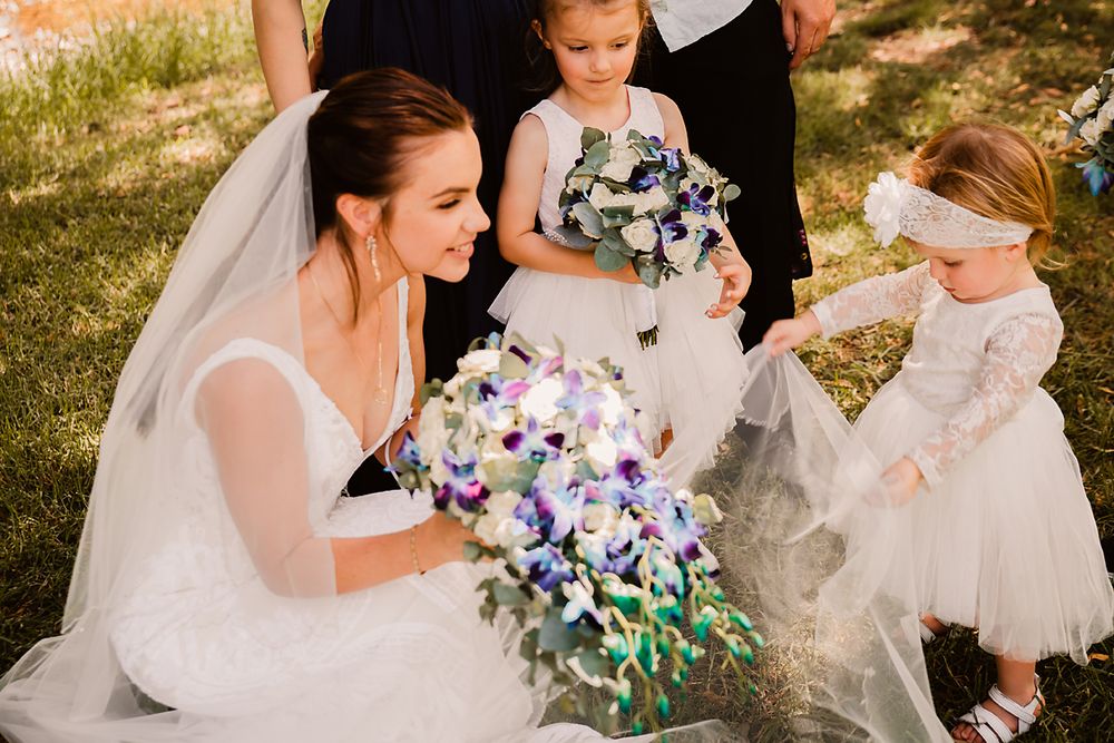 A bride in a white gown sits on grass holding a purple and blue floral bouquet while young flower girls gather around.