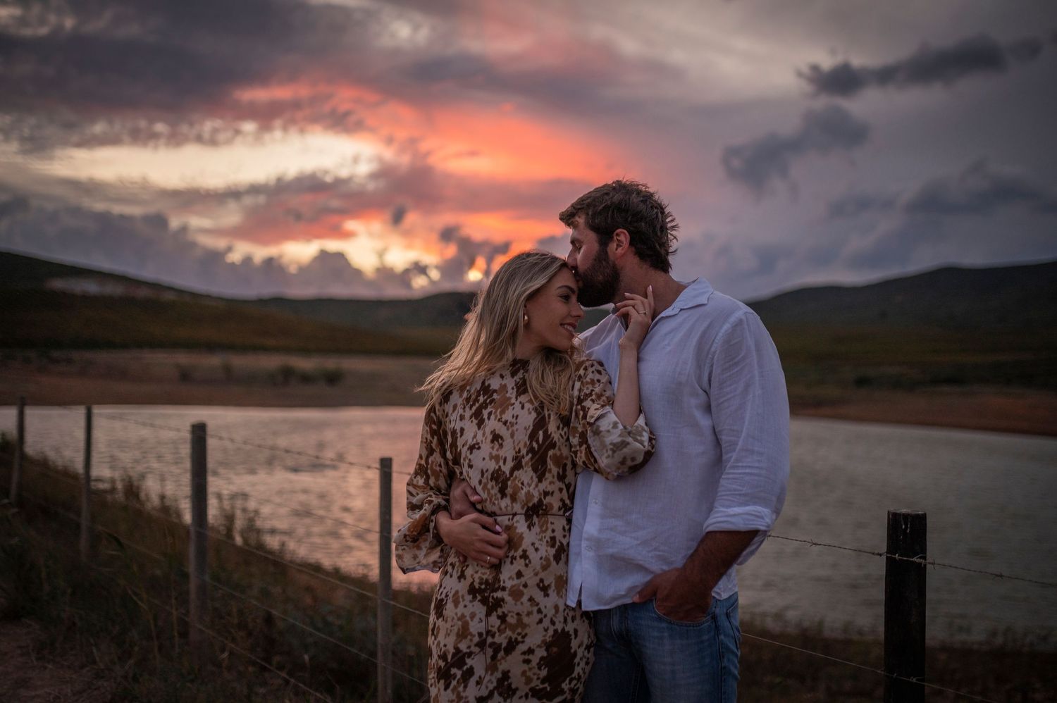 A couple embraces at sunset against a dramatic sky, with a rustic country fence and fields in the background.