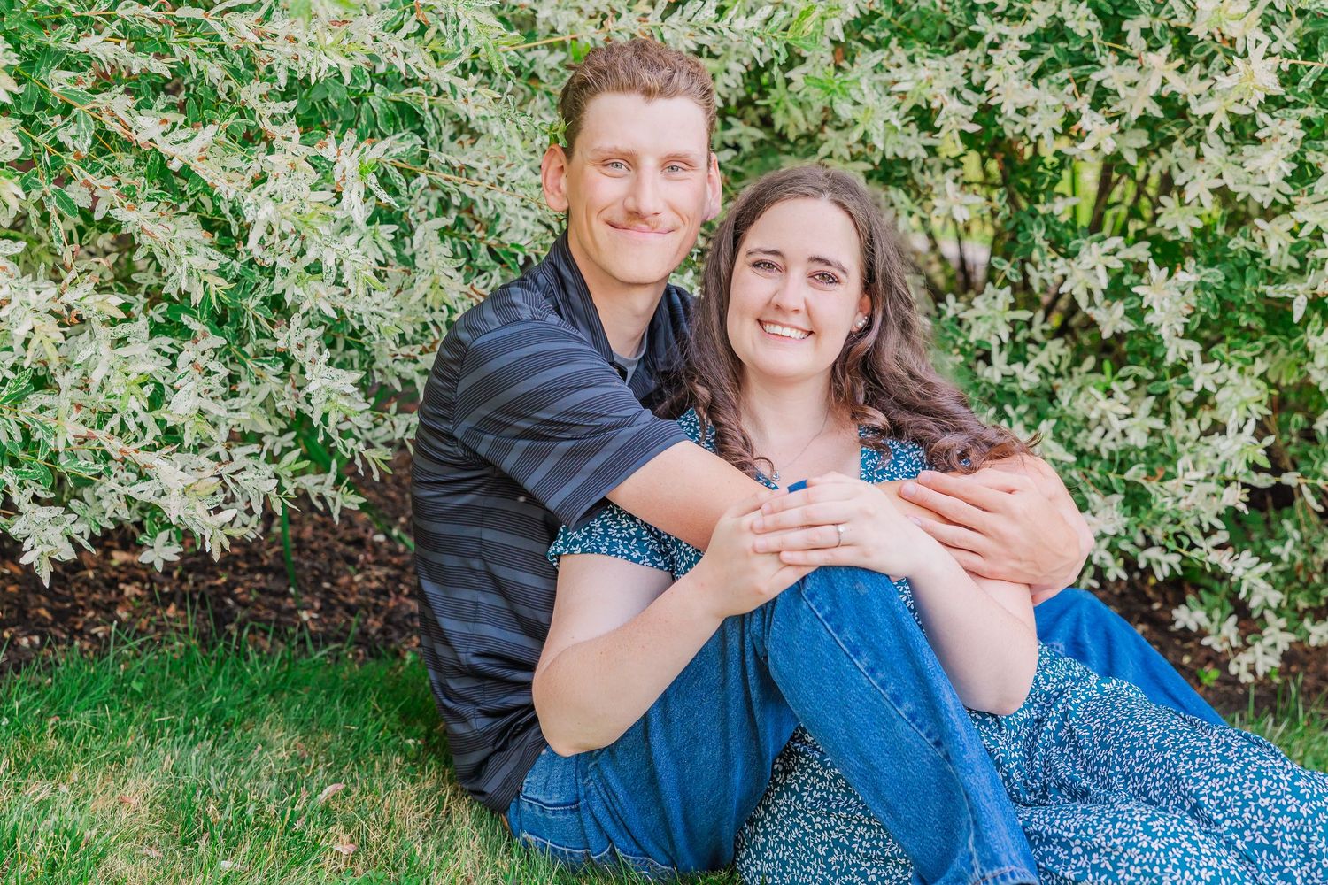 A couple in blue clothing embraces on green grass near flowering bushes in springtime.