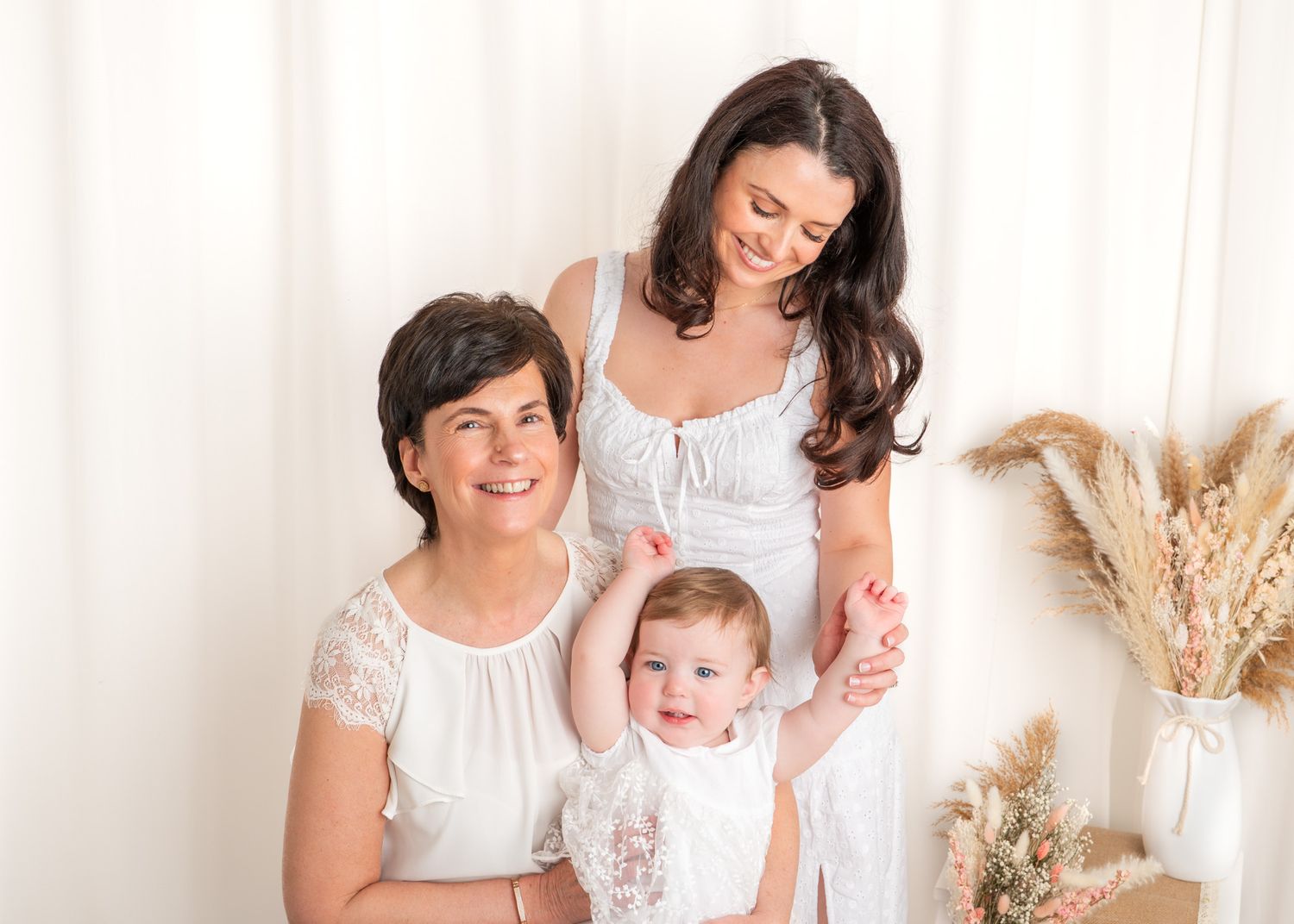 A group sitting together in a white room decorated with dried pampas grass and neutral decor during a Mother's Day photography session.