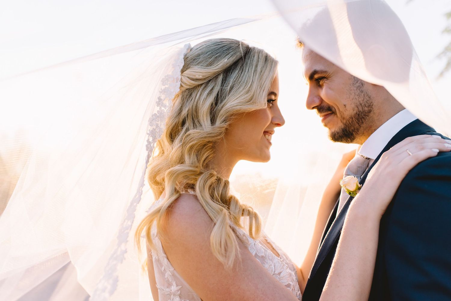 Romantic wedding couple embraces in soft glowing sunlight against translucent white curtains during sunset.