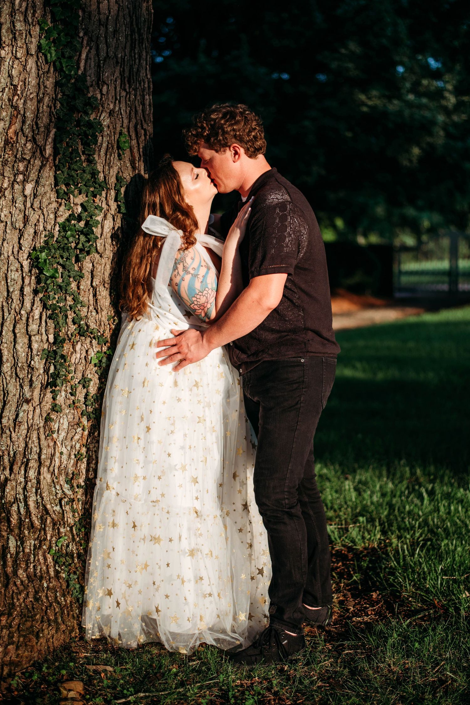 A couple shares a romantic moment beside a large tree trunk in sunlit woods. Engagement photos in VA.