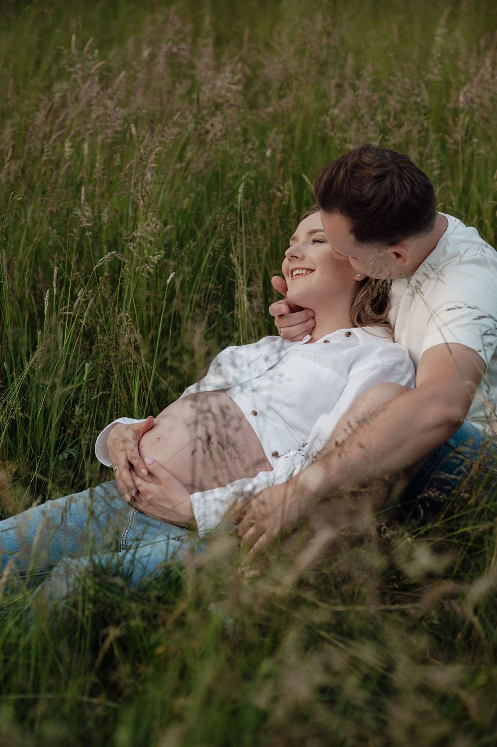 A couple relaxing together in tall grass during an outdoor maternity photoshoot.